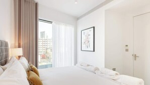 A hotel room with a large bed, a bedside table, a lamp, a framed picture on the wall, and a door to the right. - Urban Minimalist Apartment with Private Balcony (Greater London)