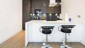 A modern kitchen with dark cabinetry, a white island with two bar stools, stainless steel appliances, and a glass backsplash. - Urban Minimalist Apartment with Private Balcony (Greater London)