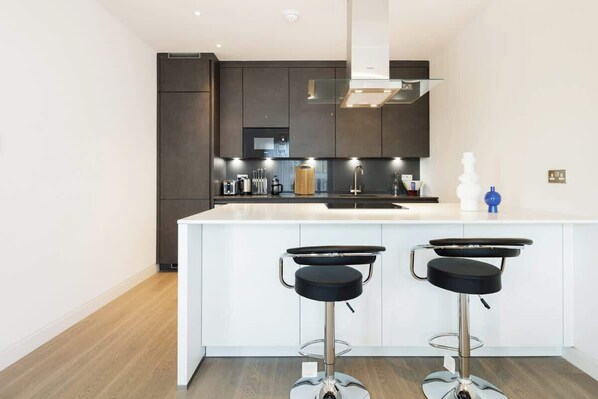 A modern kitchen with dark cabinetry, a white island with two bar stools, stainless steel appliances, and a glass backsplash. - Urban Minimalist Apartment with Private Balcony (Greater London)