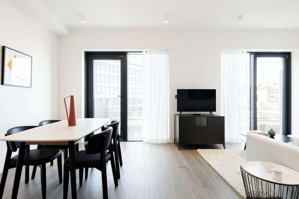 A modern living room with a dining area, a flat-screen TV, and large windows. - Urban Minimalist Apartment with Private Balcony (Greater London)