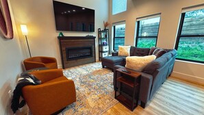 A modern living room with a fireplace, a large sofa, a smaller armchair, and a TV mounted on the wall.