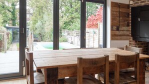 Un coin repas avec une table et des chaises en bois, de grandes portes-fenêtres coulissantes donnant sur un jardin et une cheminée en briques.