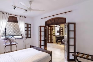 A bedroom with a bed, a desk, a chair, a ceiling fan, and an open doorway leading to another room. - Seven Pillars. 39 Galle Fort (Galle)