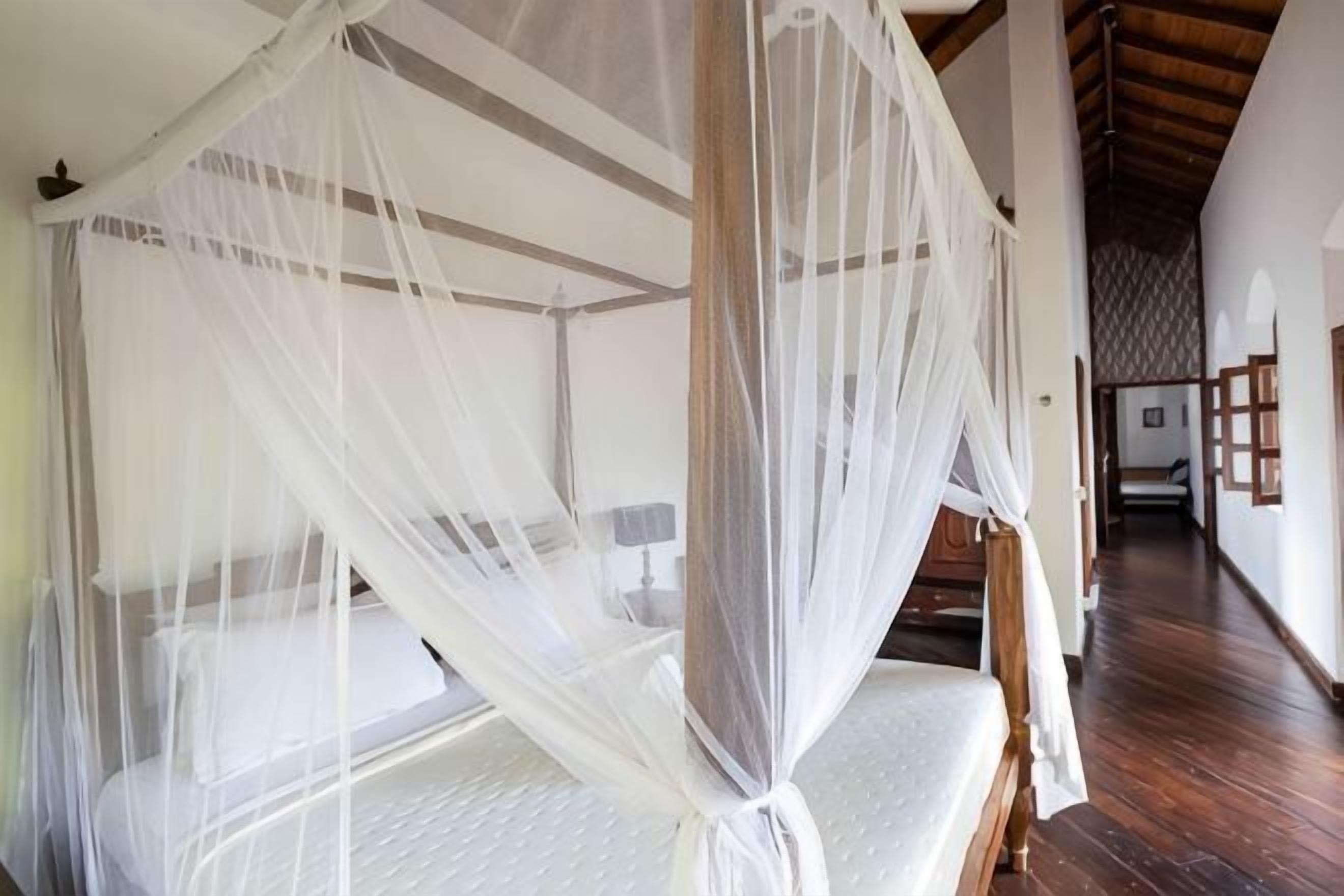 A four-poster bed with a canopy, a wooden floor, and a hallway leading to another room.