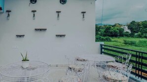 A balcony with white metal chairs and tables, a potted plant, and a view of greenery. - Hoztel Jollygrrant Doon (Dehradun)