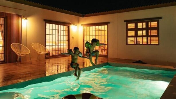 Zona de piscina con deck de madera, sillas y grandes ventanales con iluminación cálida.