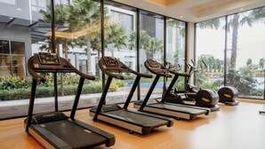 Une salle de sport équipée de tapis de course et d’appareils d’exercice, de grandes fenêtres offrant une vue sur des palmiers et un immeuble.