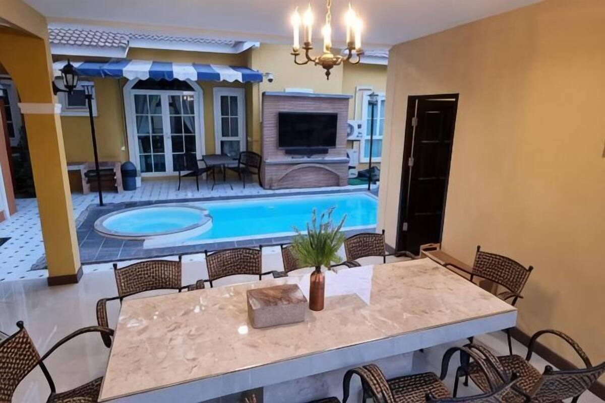 Un espace repas aménagé au bord de la piscine, avec une table en marbre, des chaises en osier, une plante en pot et un téléviseur.
