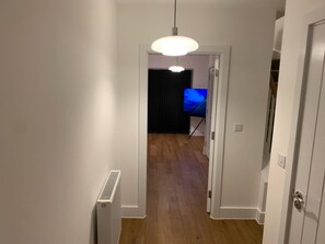 Un couloir avec un radiateur, un luminaire au plafond et une porte donnant sur une pièce où se trouve un téléviseur.