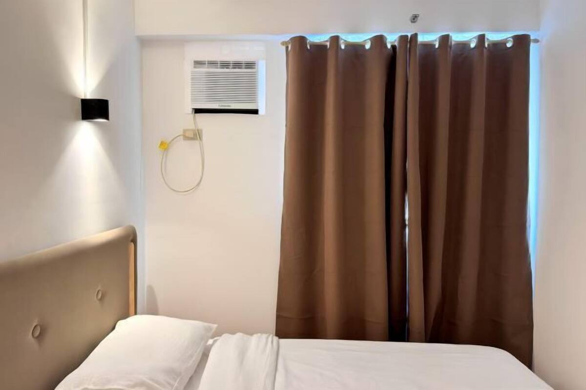 A bedroom with a bed, curtains, an air conditioning unit, and a wall-mounted light fixture.