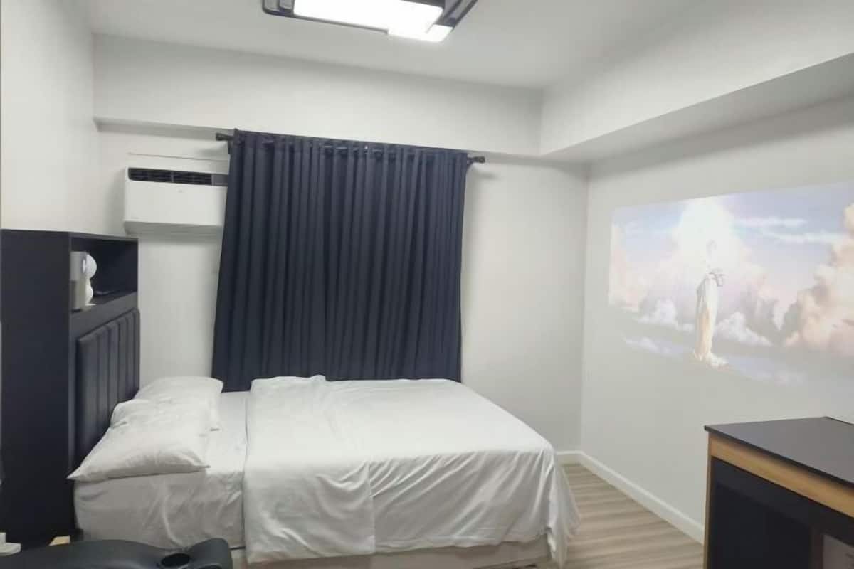 A bedroom with a bed, a dark-colored headboard, a ceiling light, a dark curtain, a wall-mounted air conditioner, and a projector screen displaying a landscape image.