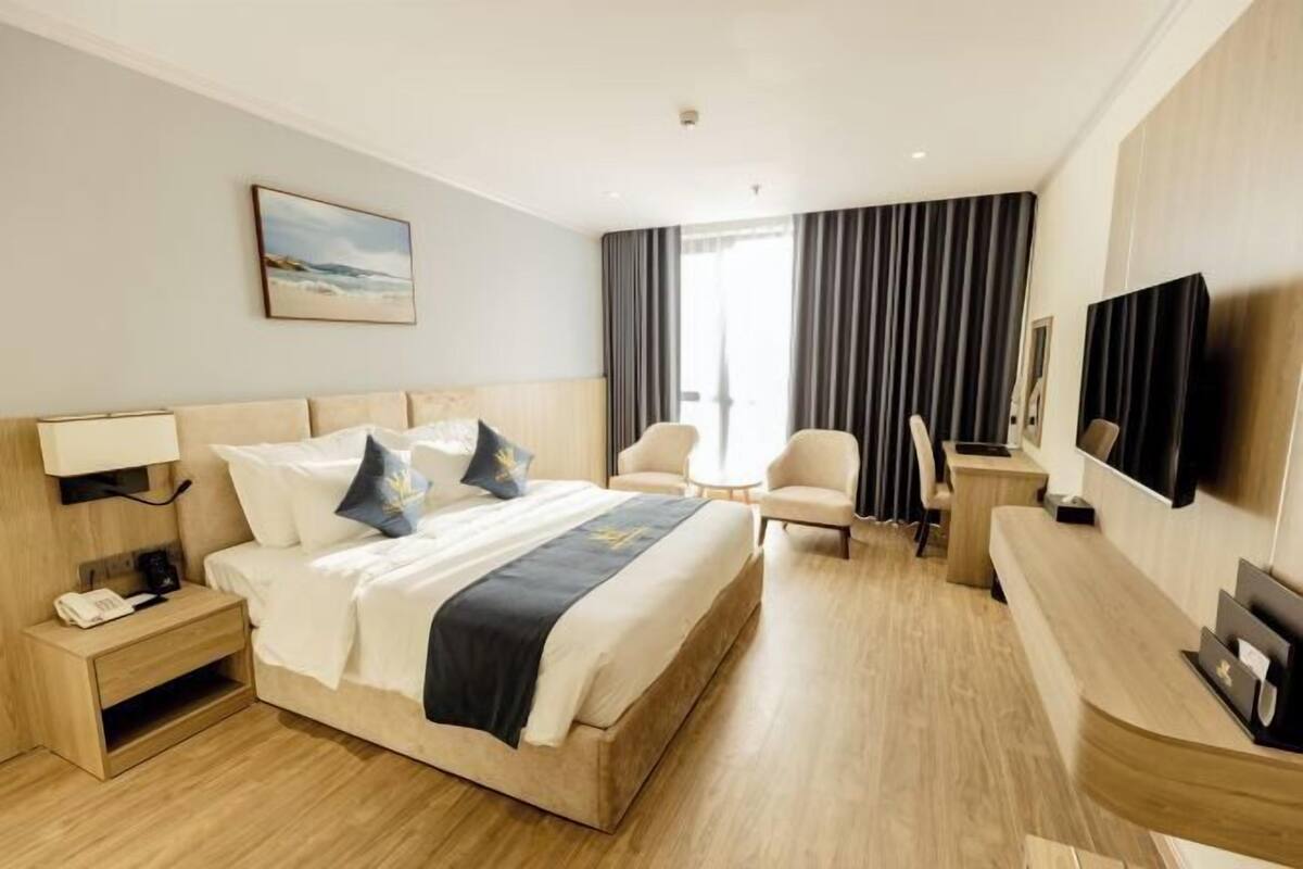 Ein Hotelzimmer mit Bett, Nachttisch, Lampe, Sessel, Schreibtisch, Fernseher und Vorhängen.