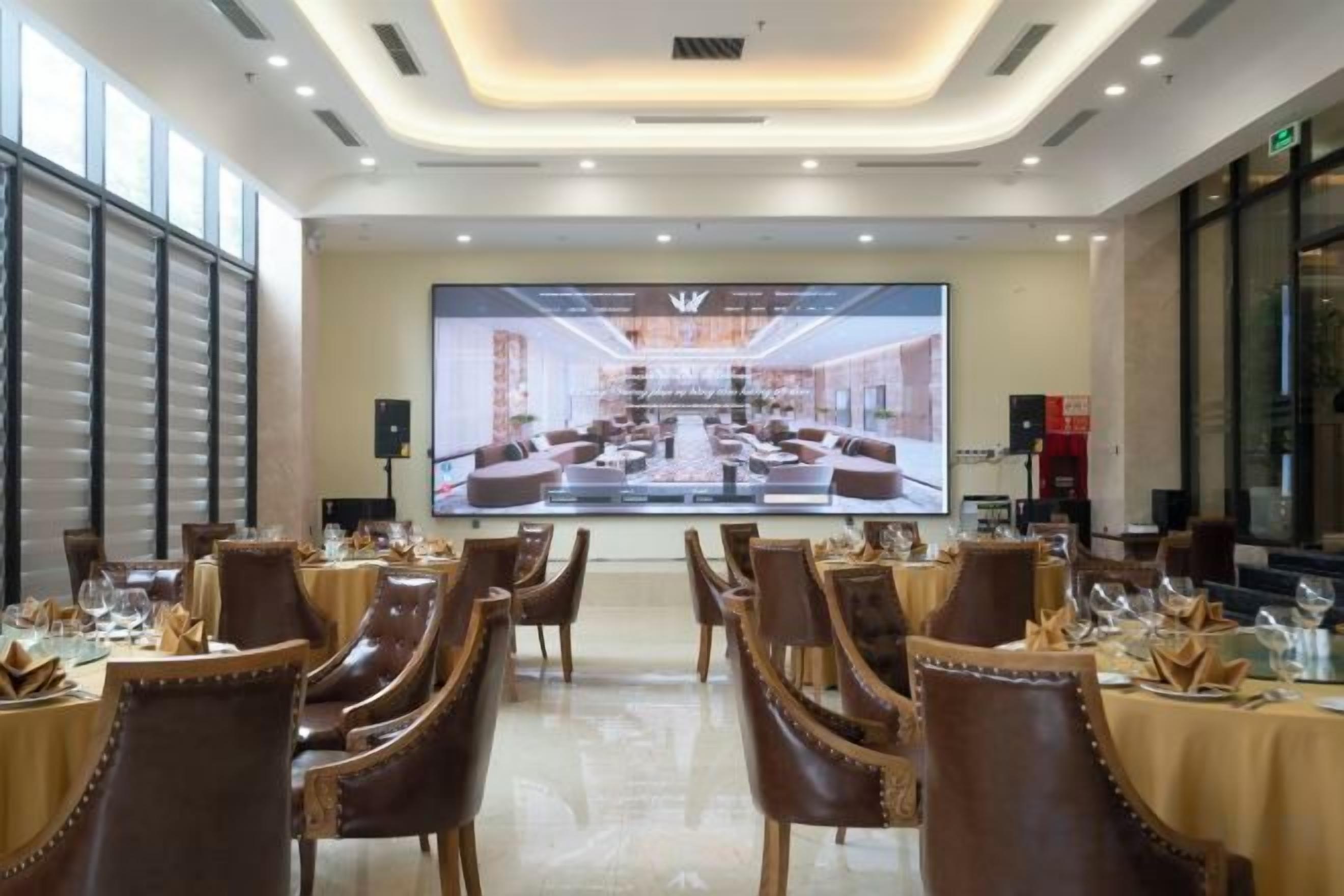 Un salón de conferencias con mesas dispuestas para una reunión, una pantalla grande que muestra la distribución de un espacio y un diseño de interiores moderno.
