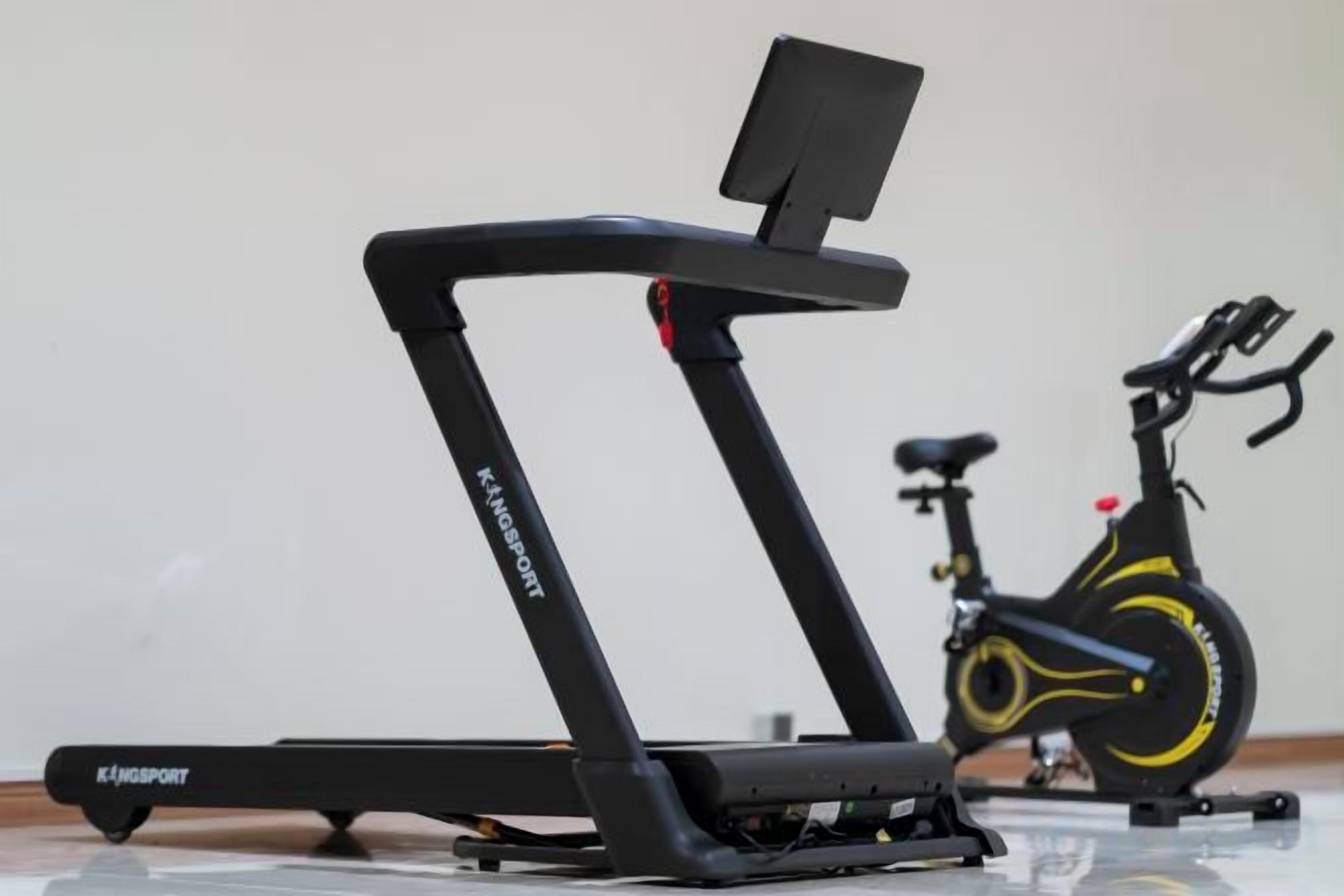 Een loopband en een hometrainer in een ruimte met een effen muur en een houten vloer.