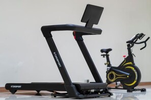 A treadmill and a stationary bike in a room with a plain wall and a wooden floor. - Hung Vuong Hotel (Mong Cai)