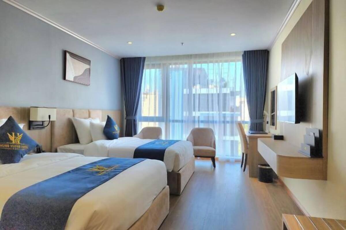 Ein Hotelzimmer mit zwei Betten, einem Schreibtisch, einem Stuhl, einem Fernseher und einem großen Fenster mit Vorhängen.