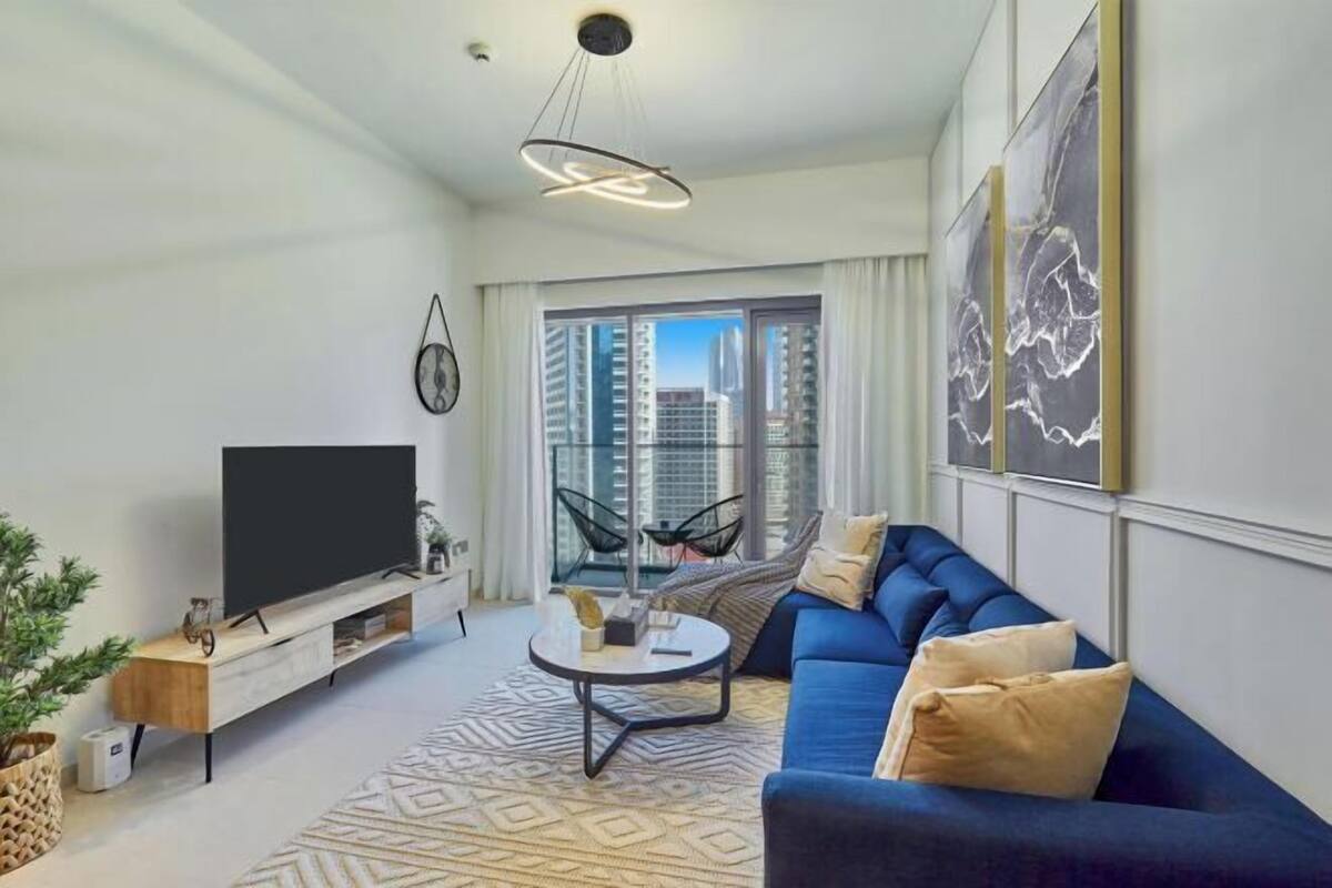 A modern living room with a large flat-screen TV, a blue sofa, a coffee table, and a view of high-rise buildings through large windows.