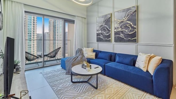 A modern living room with a blue sofa, a round coffee table, and a view of high-rise buildings through large windows. - bnbme Chic Apt w/ Balcony, 5 min to Burj Khalifa (Dubai)