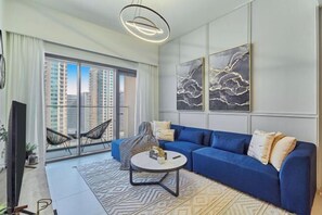 A modern living room with a blue sofa, a round coffee table, and a view of high-rise buildings through large windows. - bnbme Chic Apt w/ Balcony, 5 min to Burj Khalifa (Dubai)