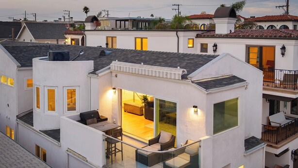 Un edificio residencial moderno de varios pisos, con balcón de cristal, zona de comedor con asientos al aire libre y un interior visible con sofá y mesa.