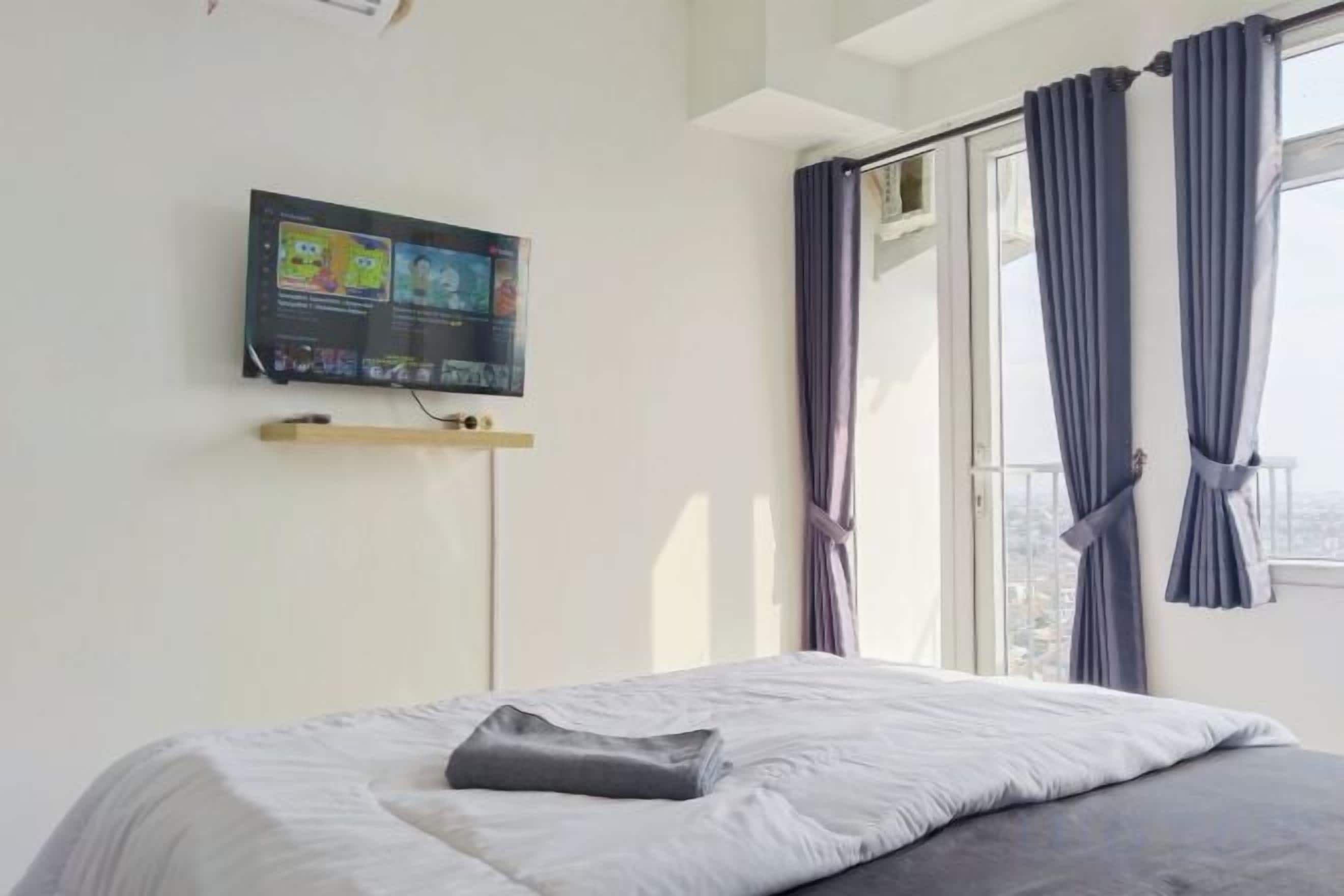 Ein Schlafzimmer mit einem Bett, einem an der Wand befestigten Fernseher und einem Fenster mit Vorhängen.