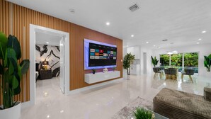 A modern living room with a large flat-screen TV mounted on a wooden panel wall, a glass coffee table, and a seating area with a cushioned bench.