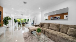 A modern living room with a large sofa, a glass coffee table, and a large mirror on the wall. There are potted plants and a dining area in the background.