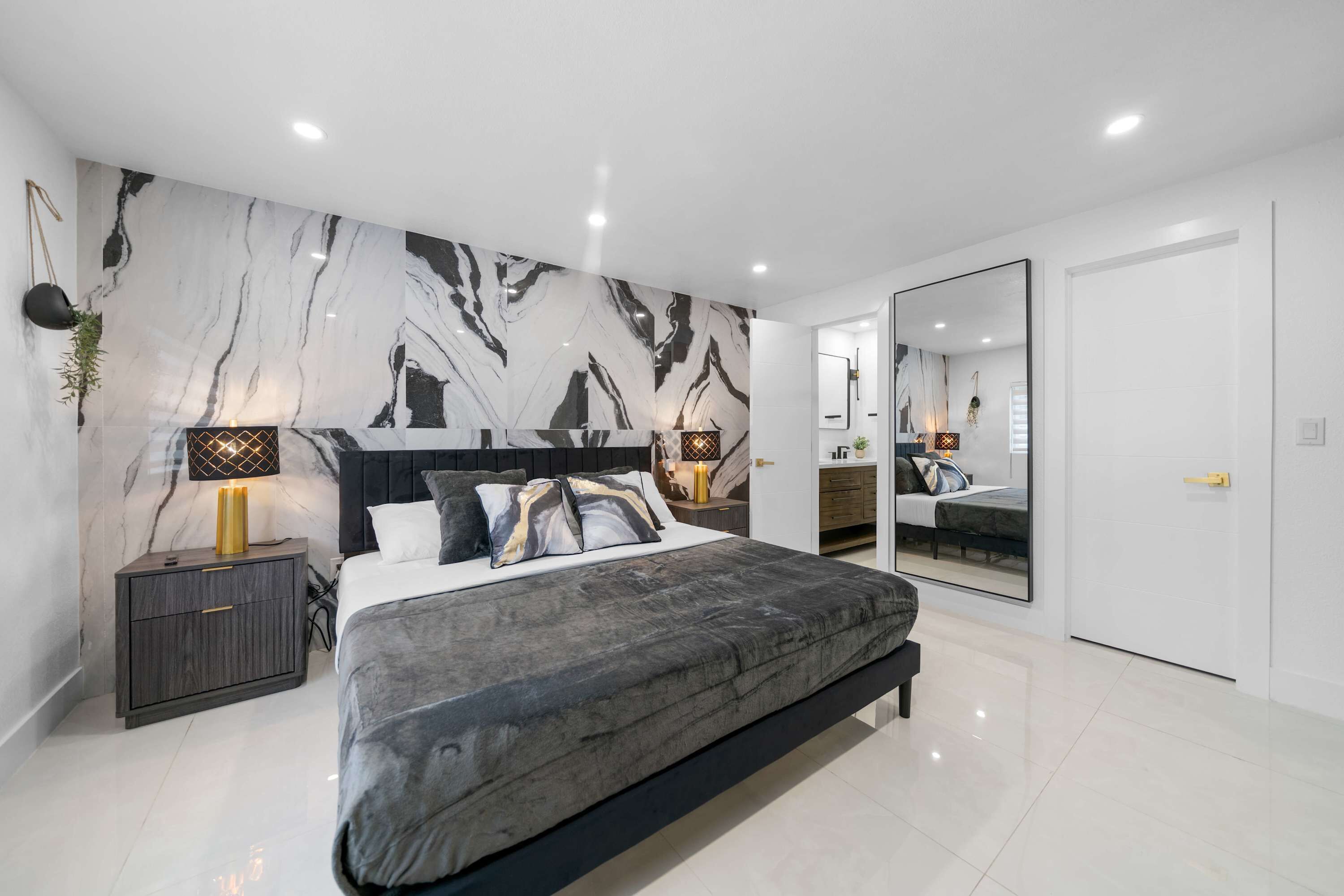 A modern bedroom with a large bed, marble-patterned wall, bedside lamps, and a full-length mirror.