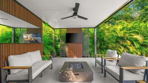 A modern outdoor seating area with a flat-screen TV, a ceiling fan, and a view of lush greenery.
