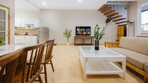 A modern living room with a wooden dining table, chairs, a glass coffee table, and a sofa. There is a kitchen area with white cabinets and a TV in the background. - VietHOME - 22 Pho Hue - Duplex 3 4 (Hanoi)