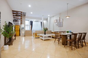 Un salón amplio con un sofá, una mesa de centro, una mesa de comedor y sillas. Se ve una escalera al fondo.