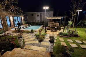 En väl upplyst uteplats med en pool, stenbelagda gångvägar och utemöbler.