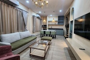 Een moderne woonkamer met een bank, voetenbankje en salontafel, een eetgedeelte met tafel en stoelen, en een keuken met kasten en een groot raam.