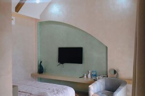 A bedroom with a bed, a chair, a television mounted on a curved wall, and a small table with items on it. - Baanjeab in khaoyai (Pak Chong)