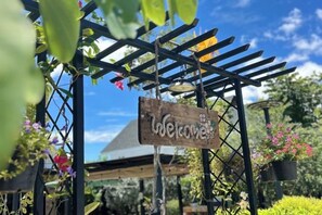 A wooden sign with the word 'Welcome' hanging from a black metal structure, surrounded by greenery and flowers. - Baanjeab in khaoyai (Pak Chong)