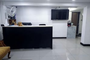 A reception area with a reception desk, a flat-screen TV mounted on the wall, a fan, and a small seating area with a chair and a sofa. - Rustic Comfort Zone (Pattaya)