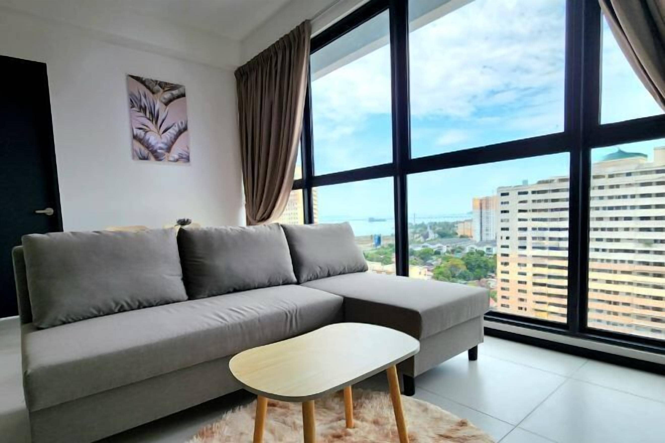 A modern living room with a grey sofa, a wooden coffee table, and a large window with a city view.
