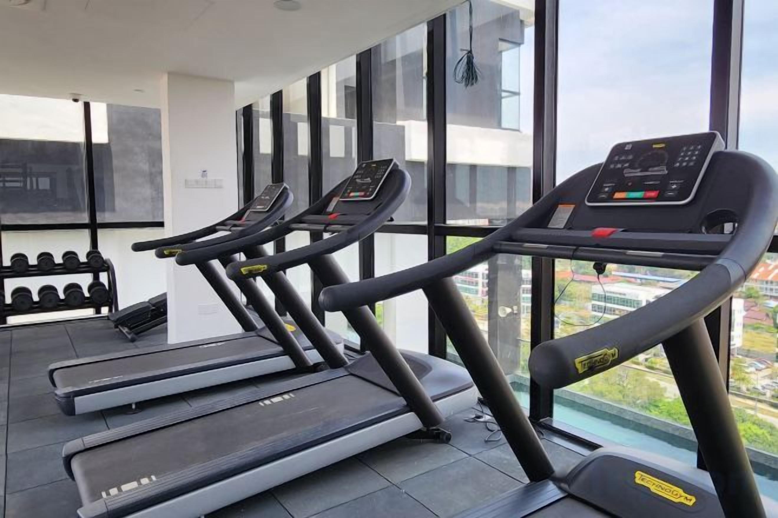 Un gimnasio con cintas de correr y mancuernas, ventanas amplias y vistas de la ciudad.