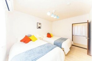 A hotel room with two beds, each with a blue blanket and colorful pillows. There is a small table with a lamp, a framed picture on the wall, and a door with curtains.