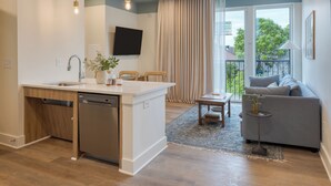 A modern kitchen with a built-in dishwasher, a dining area with a table and chairs, a gray sofa, and a large window with curtains. - 12 South Stay | 1BR Accessible Suite at Gilmore (Nashville)