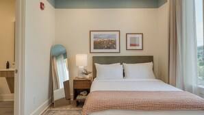 A neatly arranged hotel room with a large bed, a bedside table with a lamp, a framed picture, and a window with curtains. - 12 South Stay | 1BR Accessible Suite at Gilmore (Nashville)