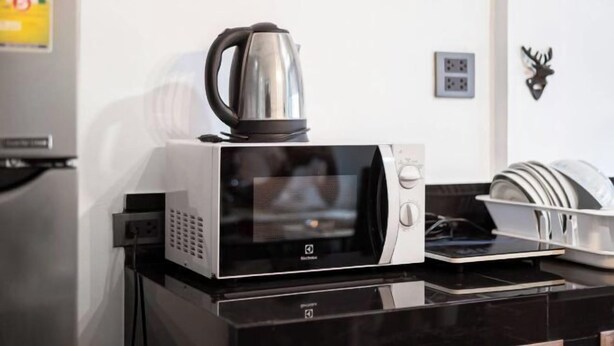 A microwave oven with a kettle on top, a dish rack with plates, and a wall-mounted electrical outlet.