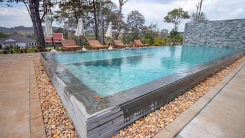 A rectangular swimming pool with a pebble-lined edge, surrounded by lounge chairs and umbrellas, set against a backdrop of trees and a stone wall.
