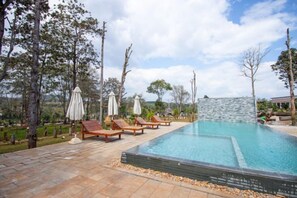 Una piscina con sillas de descanso y sombrillas, rodeada de árboles y un muro de piedra.