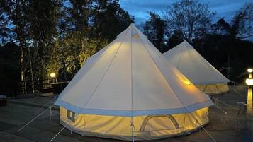 Two large, white, pyramid-shaped tents with a central light source, set on a wooden deck surrounded by trees.