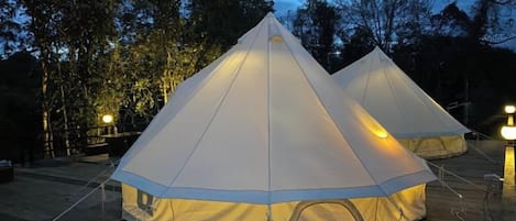 Two large, white, pyramid-shaped tents with a central light source, set on a wooden deck surrounded by trees.