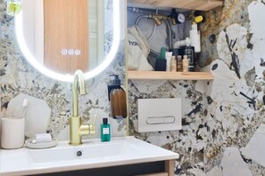 A bathroom with a white sink, a gold faucet, a mirror with a light, and a shelf with toiletries. - New! Le Vénitien, Escapade Parisienne, Sacré-Cœur (Paris)