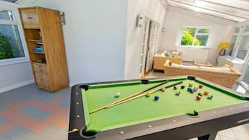 Une table de billard dans une pièce avec une fenêtre, une bibliothèque et un fauteuil en osier.