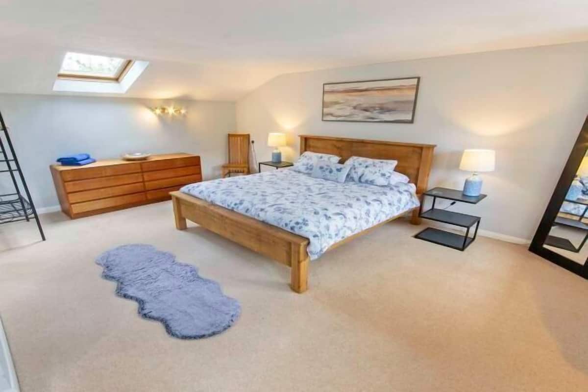 Un dormitorio con cama, cómoda, mesita de noche, lámpara, espejo, tragaluz y alfombra.