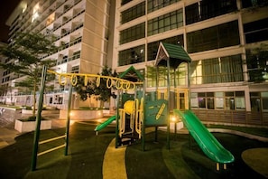 A playground with a slide and climbing structures, surrounded by a building with multiple floors and windows. - Palazio Residence By The One - Mount Austin (Tebrau)